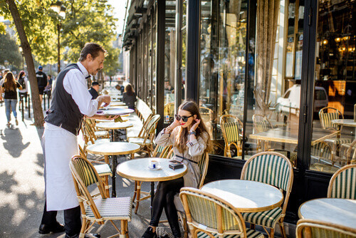 A waiter serves coffee to a young woman sitting at a traditional French Cafe in Paris.