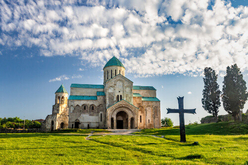 Large Christian cross standing among green grass in front of the ancient Bagrati Cathedral.