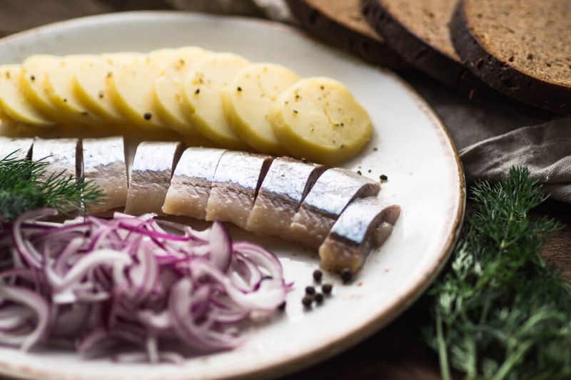 Herring fish with potato slices, rye bread, and red onion on a table.