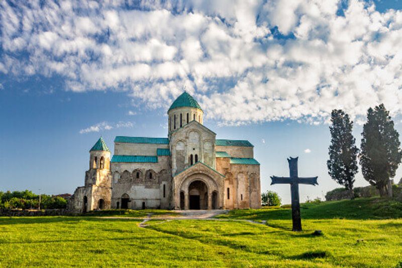 Large Christian cross standing among green grass in front of the ancient Bagrati Cathedral.