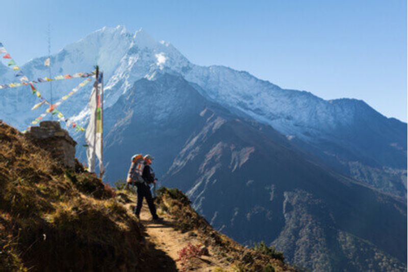 A hiker stands on a mountain edge with pack and prayer flags in Nepal.