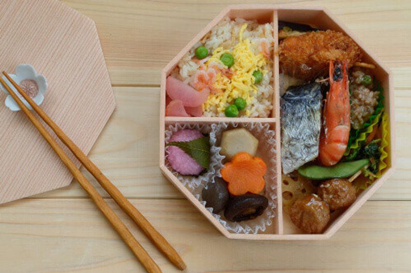 Spring inspired bento consists of sushi rice, tiger prawn, carrot, sakura mochi, croquette and shitake mushroom.