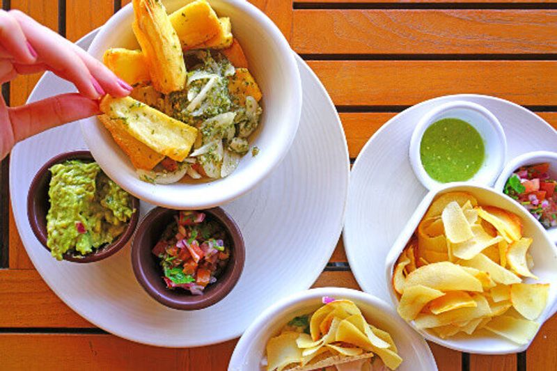 Dipping yucca fries with parsley and garlic sauce alongside side dishes of guacamole and salsa.