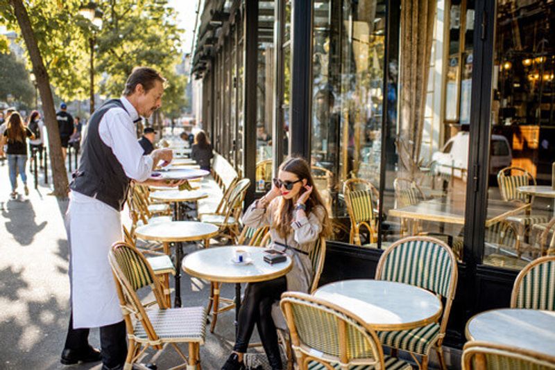 A waiter serves coffee to a young woman sitting at a traditional French Cafe in Paris.
