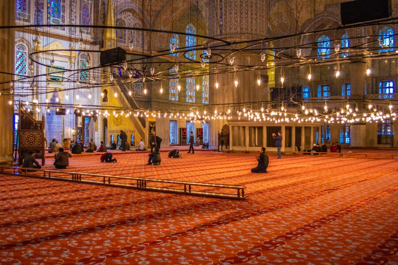 The interior of the Blue Mosque or Sultan Ahmed Mosque with red carpet and several people praying.