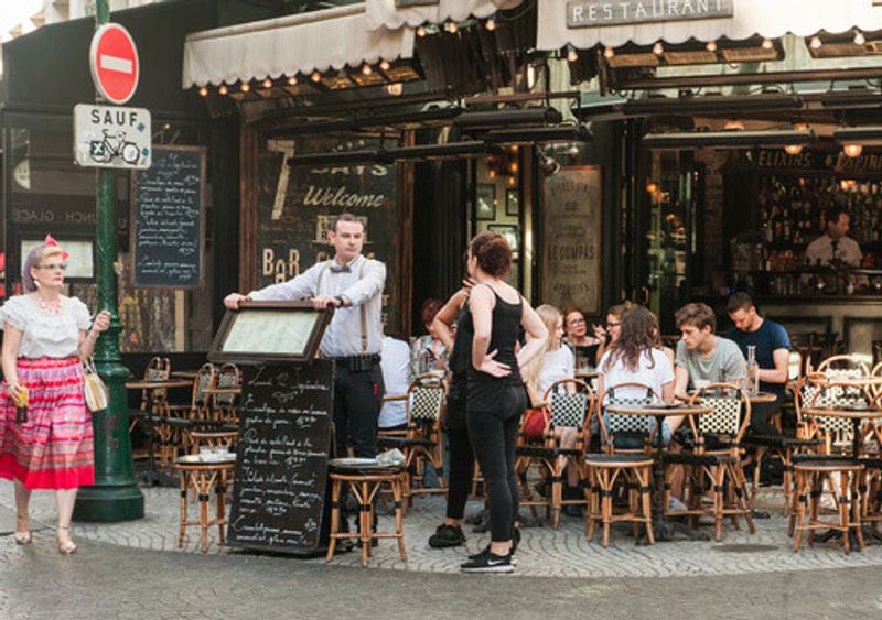 A waiter stands outside a sign in a bustling cafe in Paris, France.