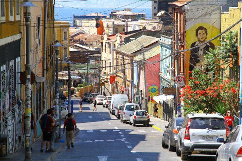 A street view of the boehmian city of Valparaiso, with artistic murals and people passing through.