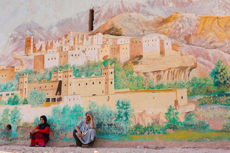 Berber locals sitting in front of the wall which has been painted with the cityscape of Erfoud.