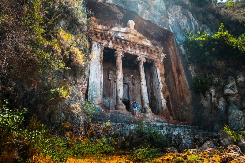 Tourist standing in front of the Amyntas Rock Tombs that are carved in steep cliff.